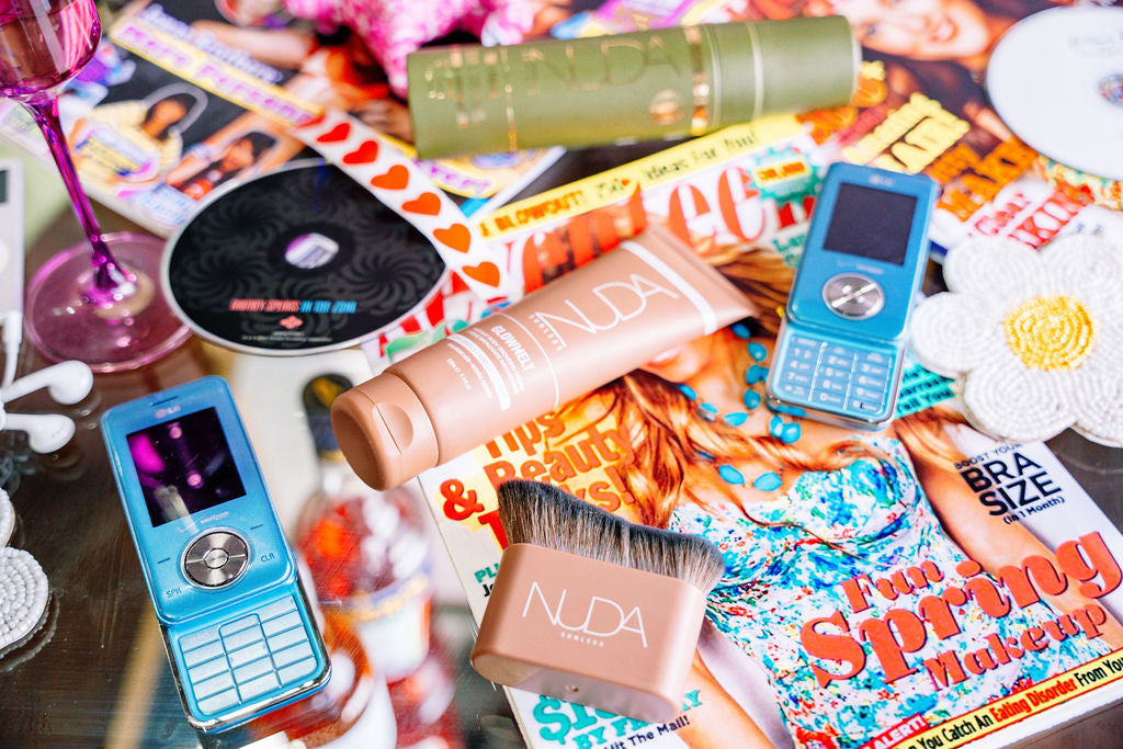 A styled flat-lay of NUDA makeup products, including a beige liquid cream, a pinkish-beige cream tube, a small blue flip phone, and a brush, arranged on top of colorful magazines.