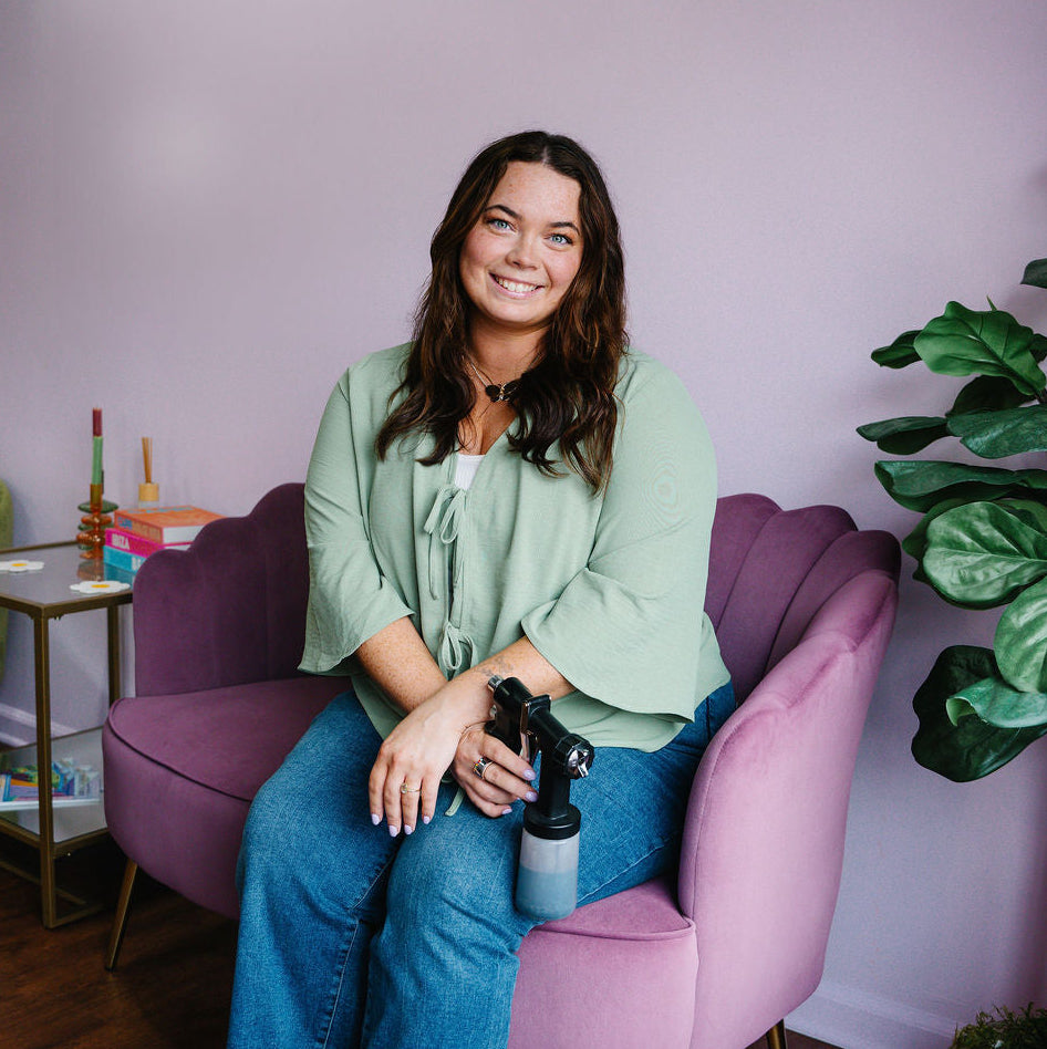 A woman with long brown hair sits smiling in a lavender armchair, wearing a sage green blouse, blue jeans, and white slide sandals. She holds a black spray bottle.