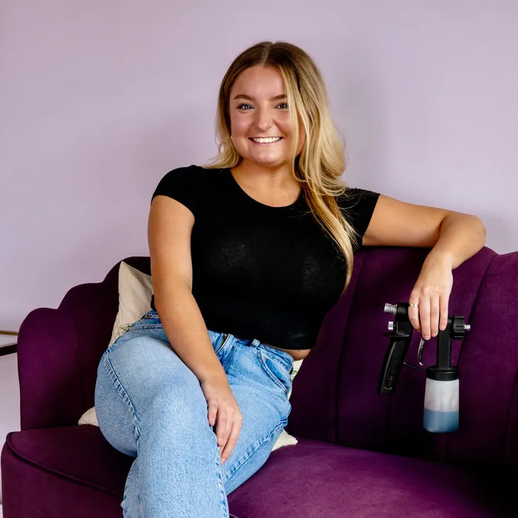 A smiling blonde woman in a black t-shirt and light blue jeans sits on a purple couch, holding a black paint spray gun with a clear container against a solid lavender wall.