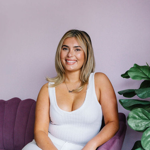 A woman with blonde hair sits smiling in a purple armchair in a room with a lavender wall and wood floors. She is wearing a white ribbed tank top and white wide-leg pants, holding a black and silver spray bottle.