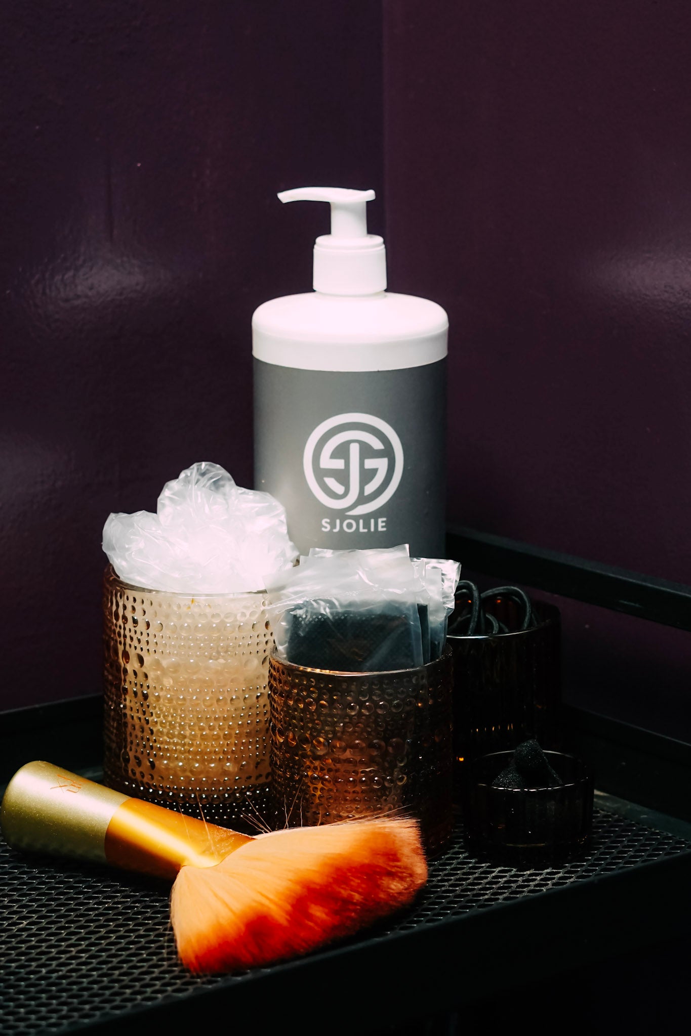 A SJOLIE branded pump bottle with a white cap and dark gray body, styled on a black tray with a gold-handled orange brush, textured holders, and a dark purple background.