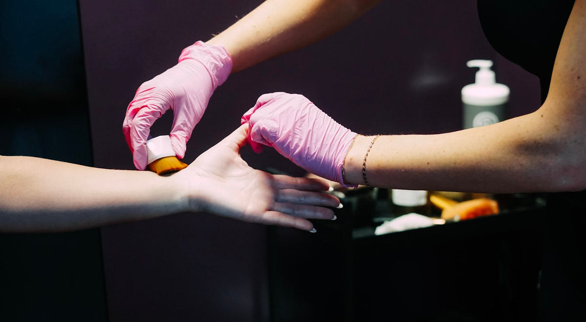 A close-up lifestyle shot of a person's hand receiving a beauty treatment, with another person's hands wearing pink gloves applying a substance. The background is a dark, modern salon setting.
