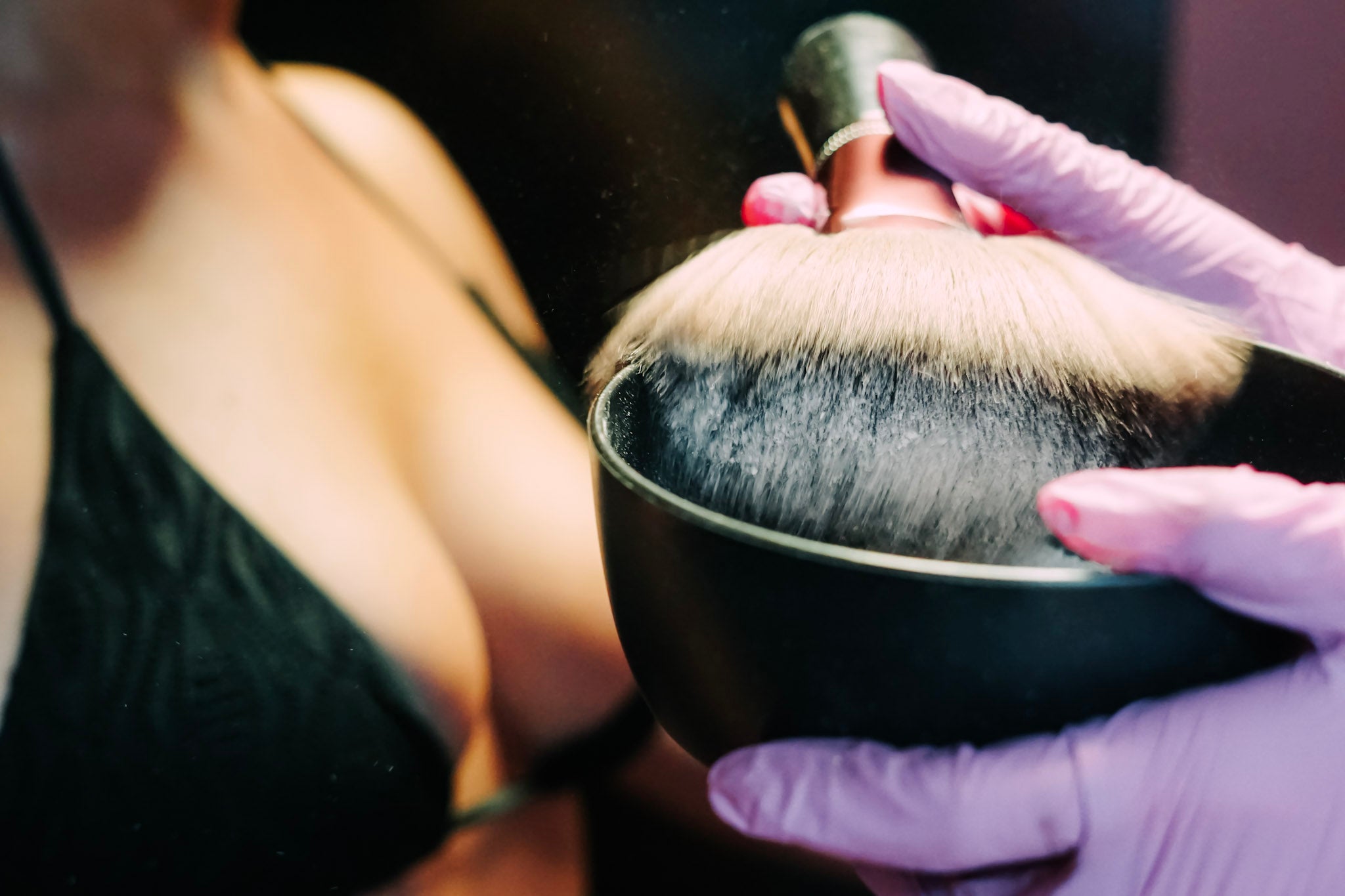 A close-up of a large makeup brush with light blonde and dark bristles being held over a black bowl by a hand wearing a lavender glove. A person in a black top is blurred in the background.